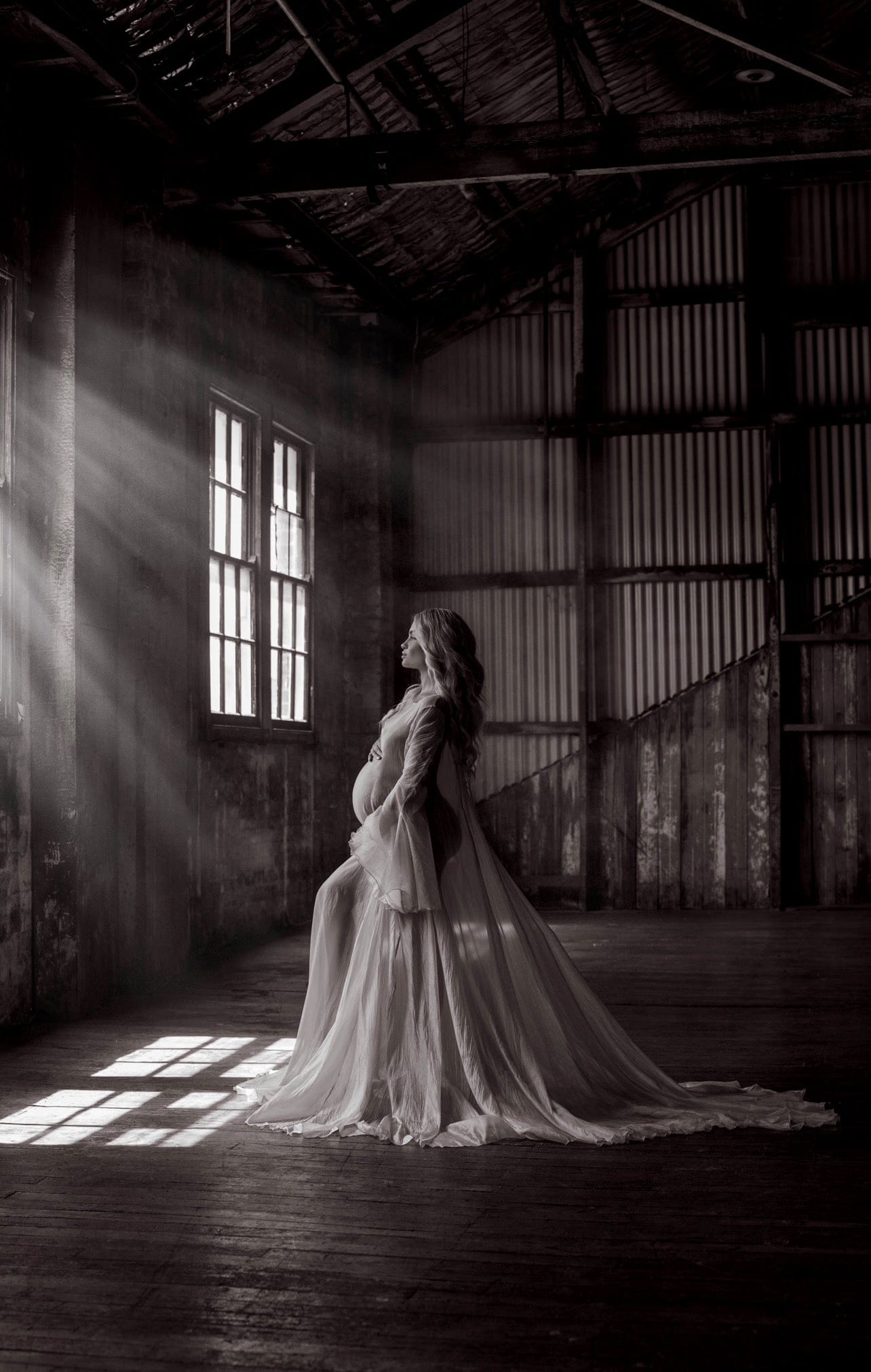 A pregnant woman wearing a long, flowy, white maternity dress from Everything Lace Hire with subtle pleats, standing in a room with natural light streaming through windows. Maternity Photoshoot Dresses Australia