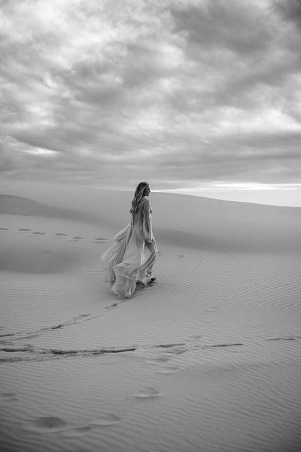 Pregnant lady in Everything Lace Hire Minna Gown a long dress walking on sand dunes under a cloudy sky. Stunning maternity photoshoot dress