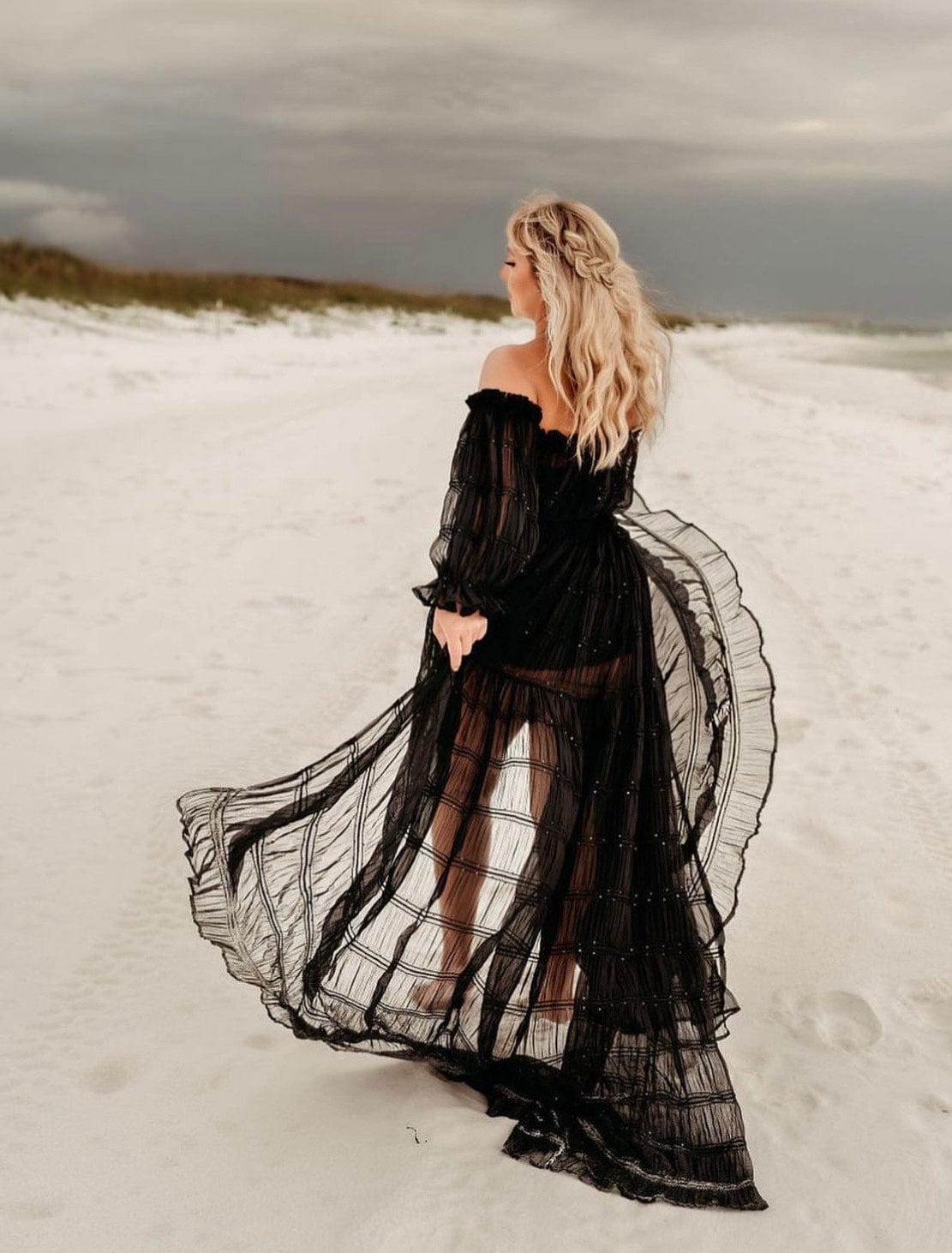 A lady stands on a beach of white sand wearing Everything Lace Hire black chiffon two piece