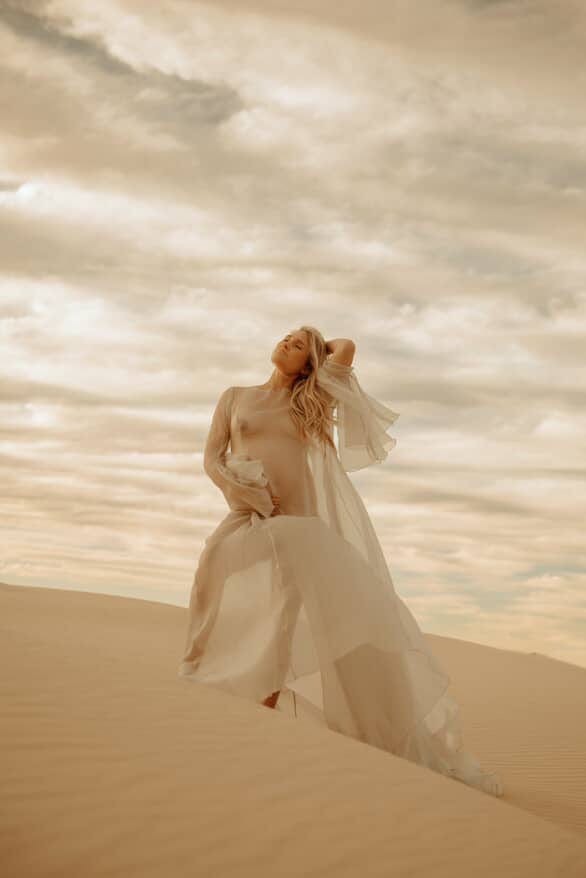 Pregnant Woman in the Minna Gown from Everything Lace Hire a flowing maternity photoshoot dress standing on sand dunes with a dramatic sky.