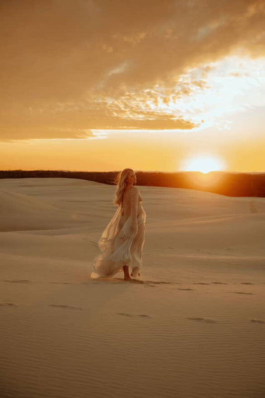 Woman in the Minna Gown a long flowing maternity photoshoot dress from Everything Lace Hire standing on sand dunes at sunset.