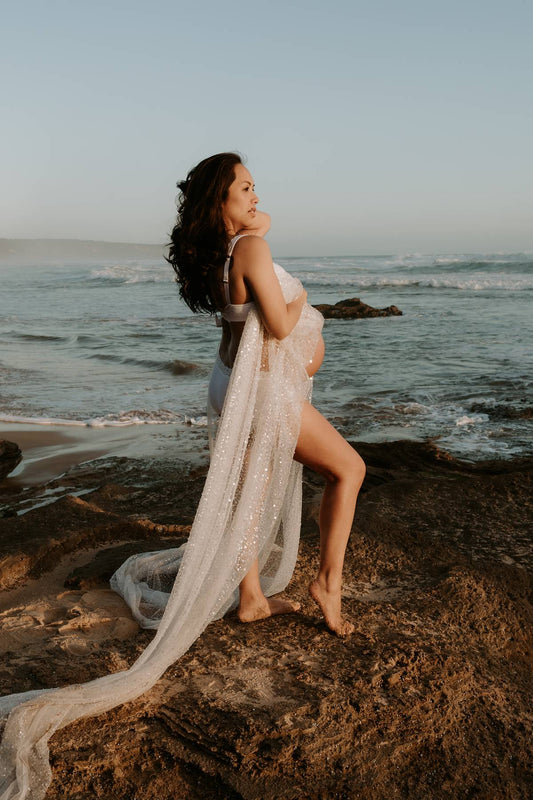 Pregnant woman wearing Everything Lace sequin beaded flowing drape standing on a rocky beach with ocean waves in the background.
