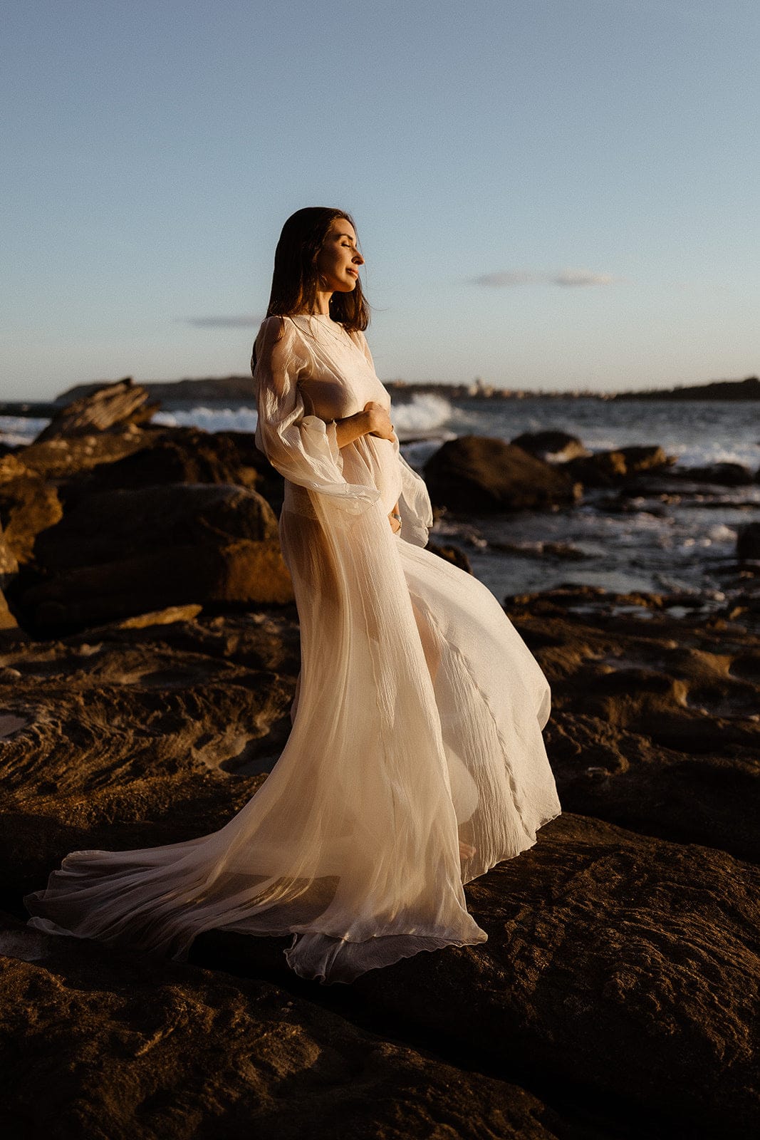 Pregnant Woman in Everything Lace Hire Vienna Gown a long flowy Maternity Photoshoot Dress standing on rocky beach at sunset capturing her bump during her maternity photoshoot