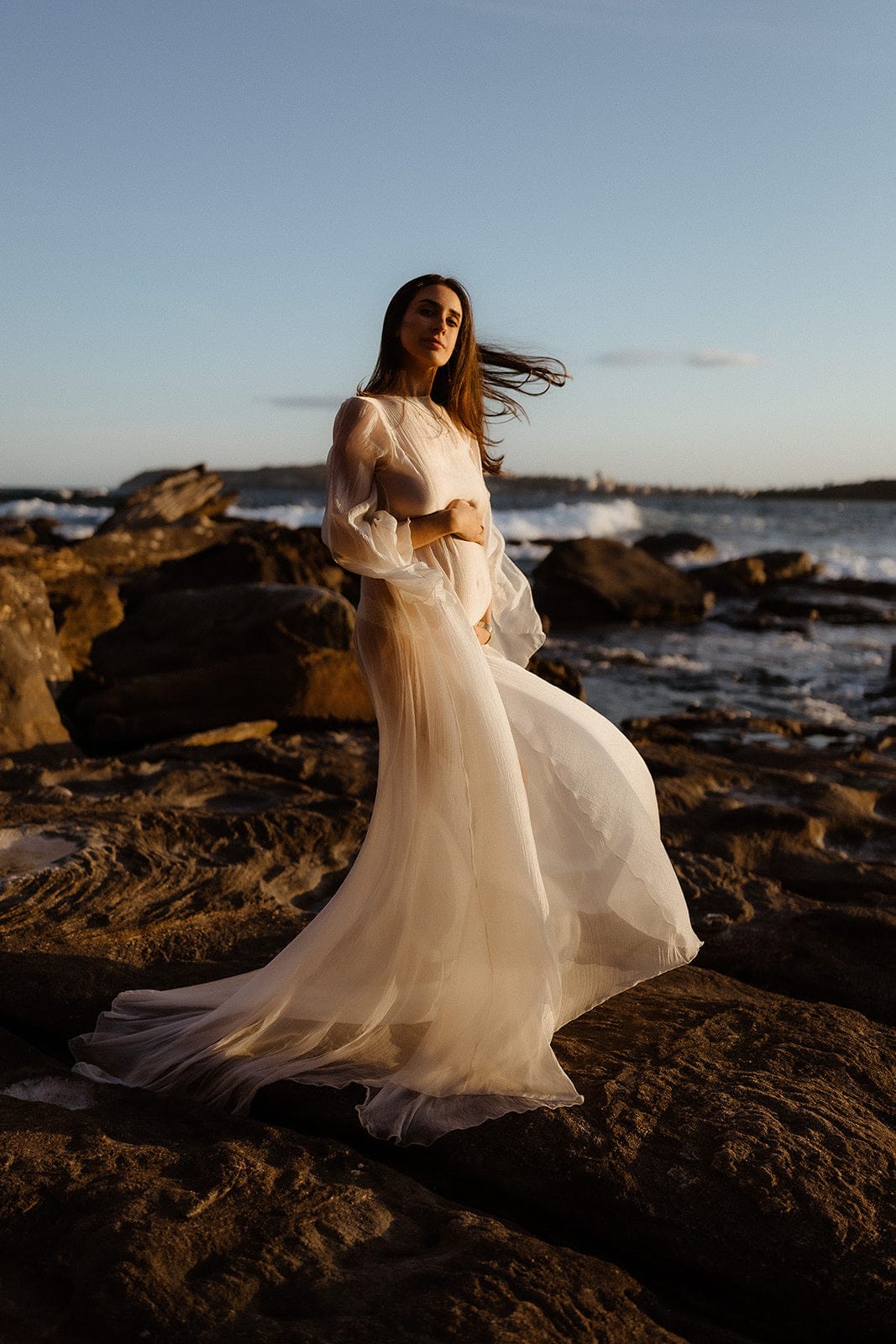 Pregnant Woman in Everything Lace Hire Vienna Gown a long flowy Maternity Photoshoot Dress standing on rocky beach at sunset with waves in the background capturing her bump during her maternity photoshoot