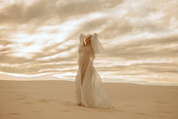 Pregnant lady in a long, light-colored dress from Everything Lace Hire standing on a sandy beach with waves in the background. The most stunning dress to wear for maternity photos