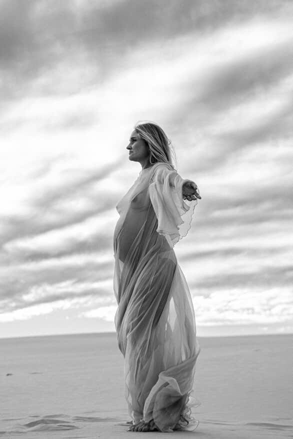 Woman in a flowing dress standing on a beach with a dramatic sky. Everything Lace Hire collection of maternity photoshoot dresses