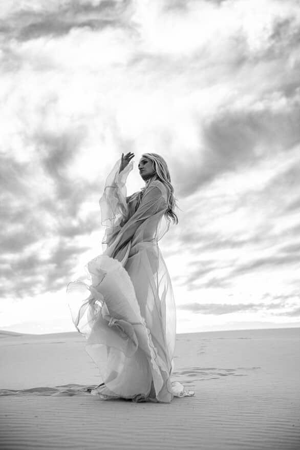 Pregnant lady in a flowing dress from Everything Lace Hire standing on sand with a dramatic sky