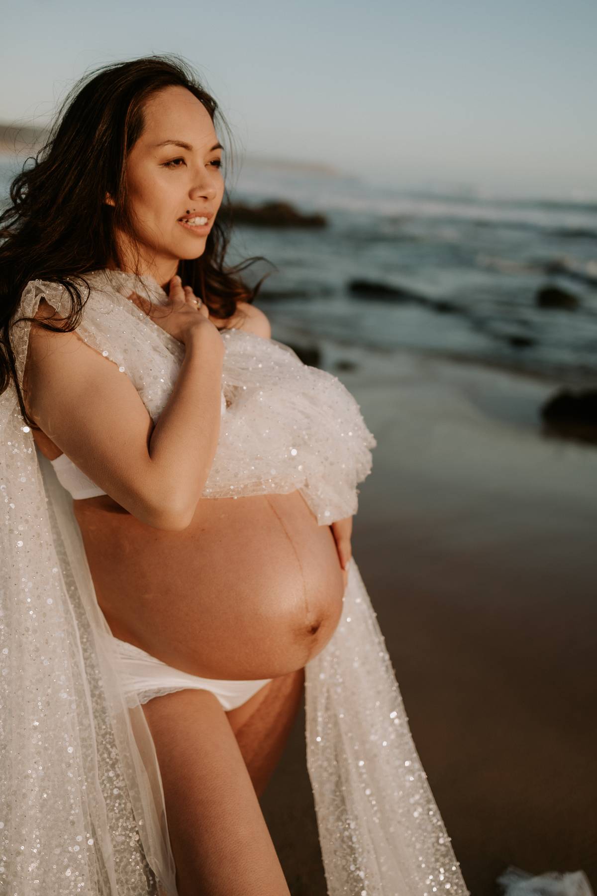 Pregnant woman wearing Everything Lace Hire sparkly maternity drape across her body to showcase her pregnancy bump standing on a beach during her maternity photoshoot.