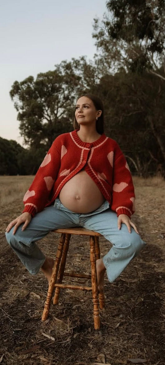 a pregnant lady sits on the chair wearing Everything Lace hire Lover Knit a thick chunky red cardigan with pink love hearts