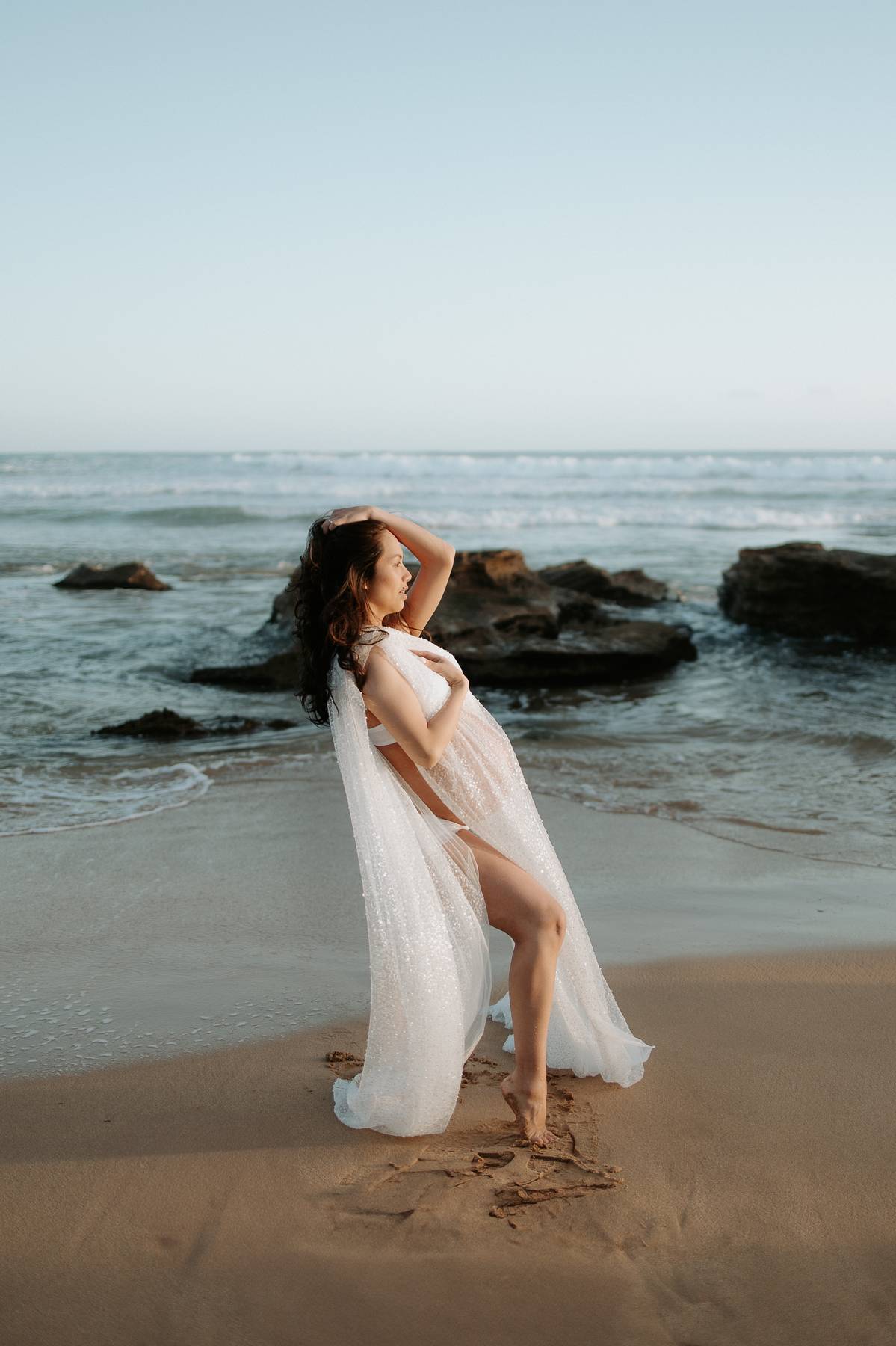 Pregnant Woman in a white maternity dress drape from Everything Lace Hire standing on a beach with ocean and rocks in the background during her beach maternity photoshoot