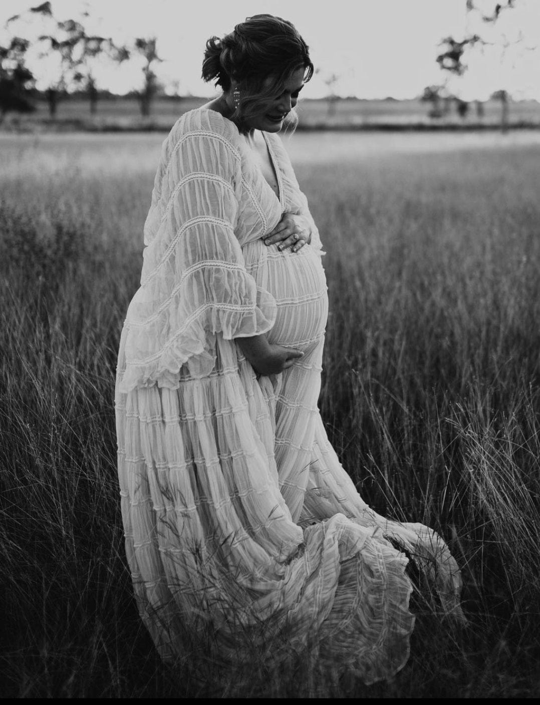 Pregnant lady standing amongst the dried grass looking down at her bump while wearing Everything Lace Hire Wonderful Moments Gown