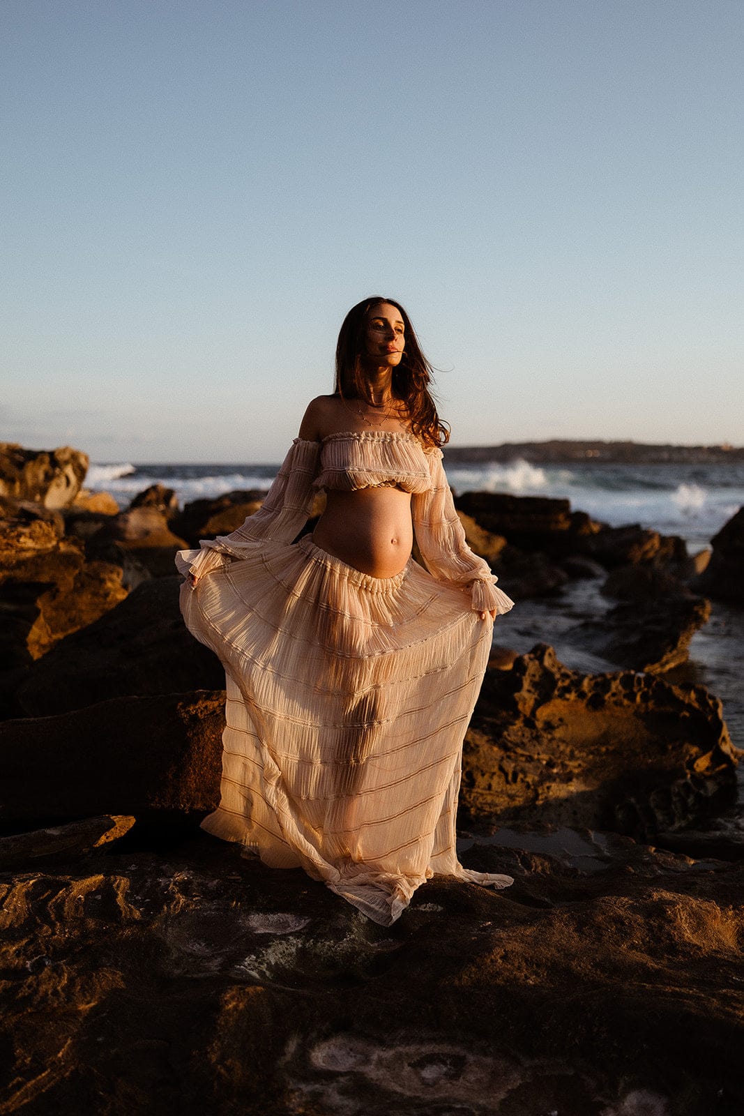 Pregnant woman in Everything Lace Hire Wonderful Moments Set a flowing two piece set in nude standing on rocky beach at sunset during her maternity photoshoot