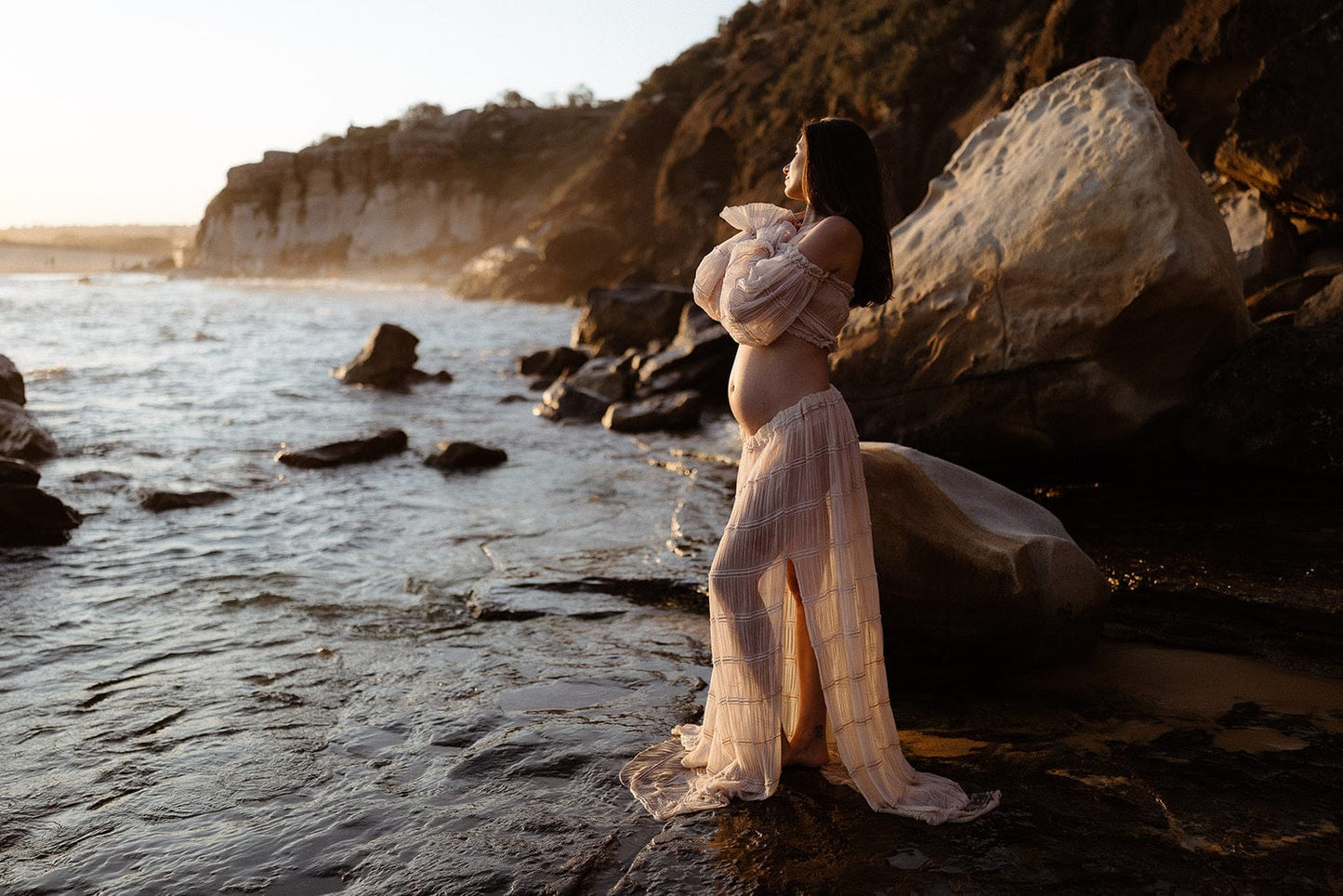 Woman in a flowing two piece set from Everything Lace Hire standing on a rocky beach at sunset