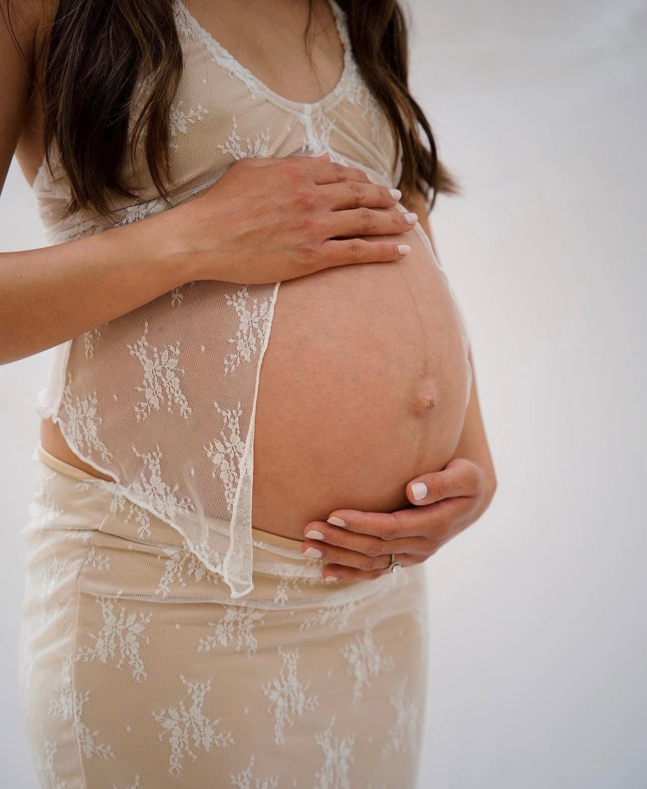 Pregnant woman wearing a lace dress  from Everything Lace Hire holding her belly against a plain background during her maternity photoshoot
