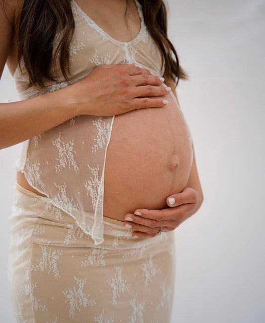Pregnant woman wearing a lace dress  from Everything Lace Hire holding her belly against a plain background during her maternity photoshoot