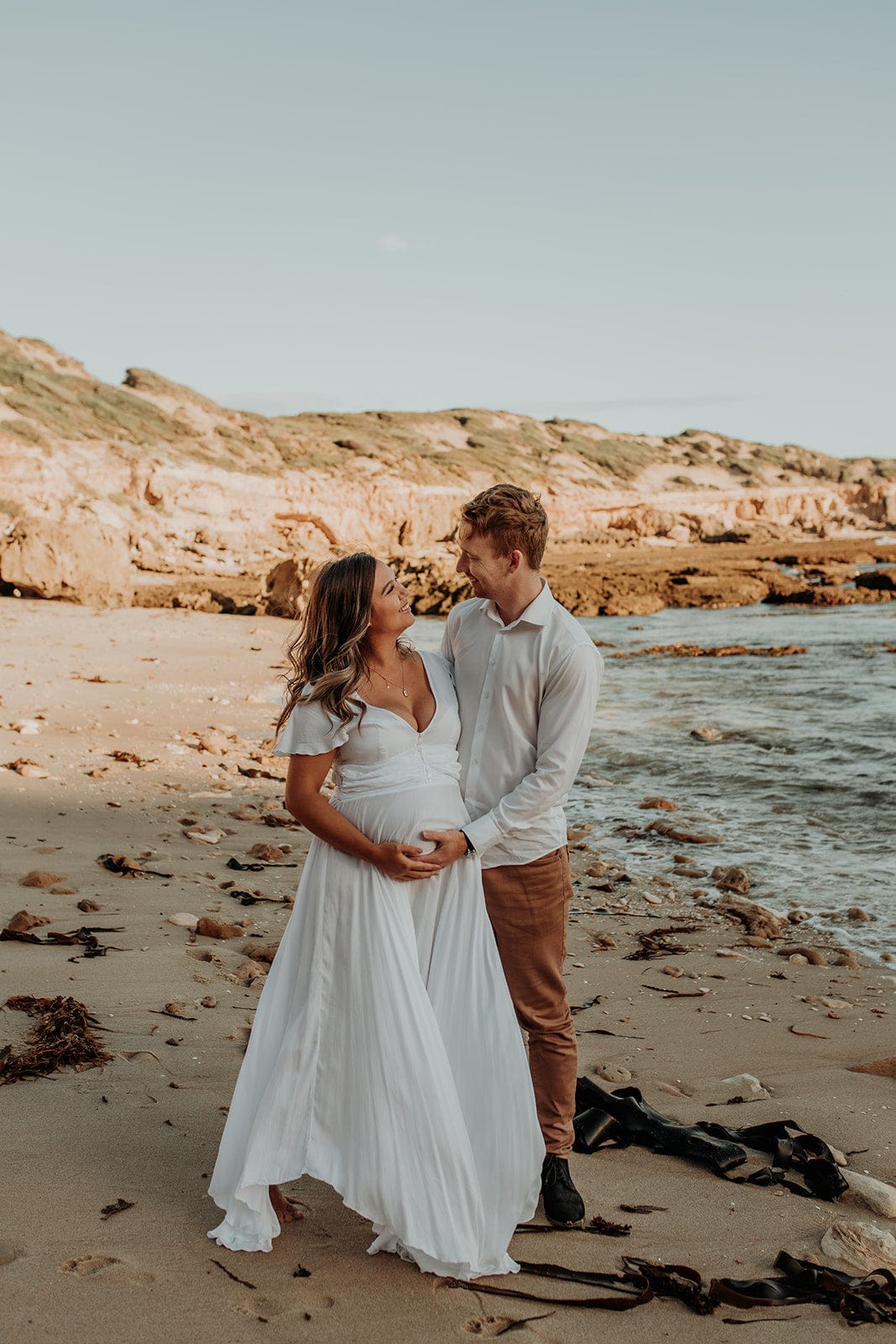 A couple takes a moment to look at each other during their maternity photoshoot on the beach in Blairgowrie. Mum is wearing a white Coven and Co Halo Dress