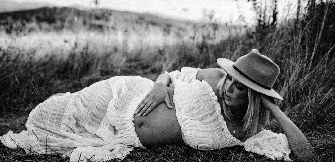 A beautiful pregnant mother lays on the grass cupping her bump wearing Everything Lace Hire's Anabelle Two Piece Set and wide brim hat.